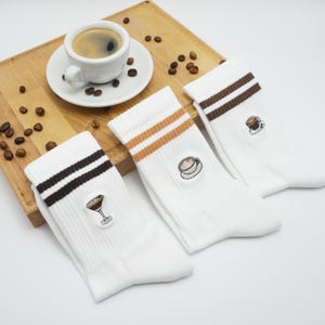 Könnte beinhalten: Weiße Socken mit braunen und beigefarbenen Streifen, jeweils mit einem gestickten Kaffee-Motiv. Eine Tasse Kaffee und Kaffeebohnen liegen auf einem Holzbrett.