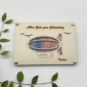 May include: A wooden card with a laser-cut design of a blimp holding a banknote. The card says "Alles Gute zum Geburtstag" and "Torsten".