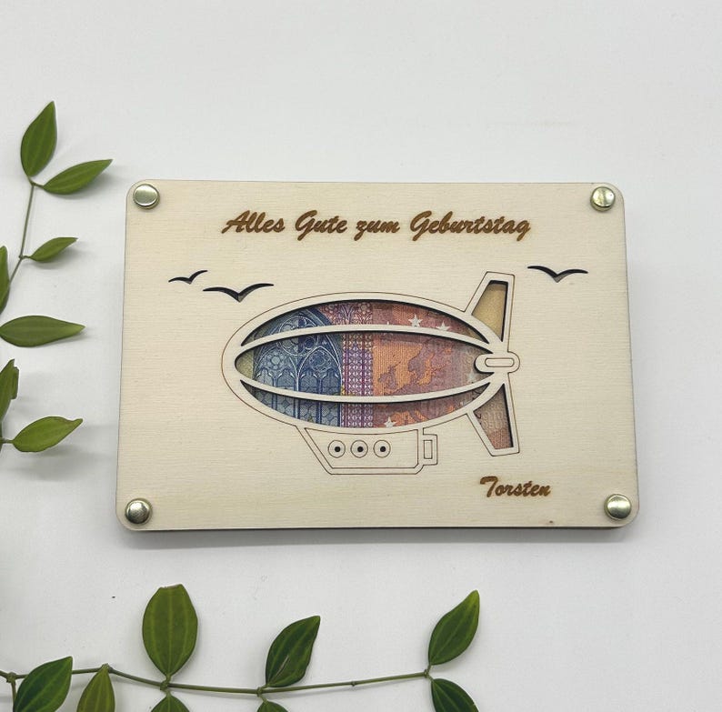 May include: A wooden card with a laser-cut design of a blimp holding a banknote. The card says "Alles Gute zum Geburtstag" and "Torsten".