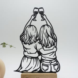 May include: Black silhouette of two figures toasting wine glasses. The figures are embracing, with their backs to the viewer. The artwork is mounted on a wooden base, suggesting a decorative item or a gift.