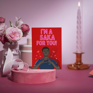 May include: A red Valentine's Day card with a cartoon illustration of a man with a heart in his hands. The card says "I'm a Saka for you!" in pink letters.
