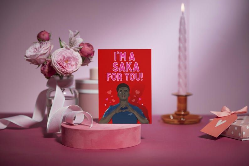 May include: A red Valentine's Day card with a cartoon illustration of a man with a heart in his hands. The card says "I'm a Saka for you!" in pink letters.
