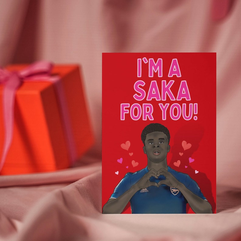 May include: A red card with the text "I'm a Saka for you!" in pink letters. A cartoon image of a soccer player making a heart shape with his hands is on the card.