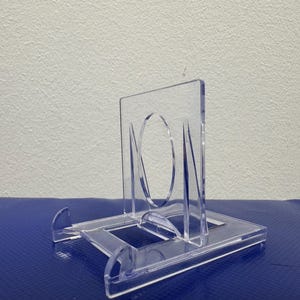 May include: A clear plastic display stand for a small object, such as a coin or a medal. The stand has a square base and a raised platform with a circular cutout in the center.