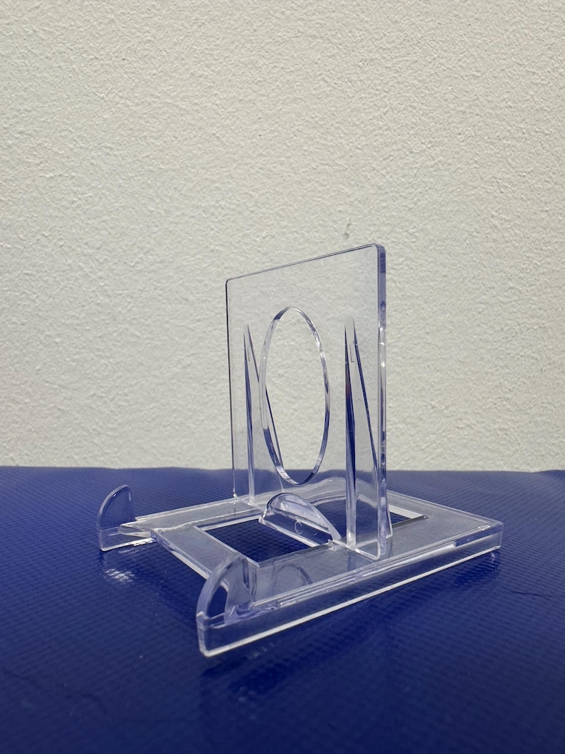 May include: A clear plastic display stand for a small object, such as a coin or a medal. The stand has a square base and a raised platform with a circular cutout in the center.