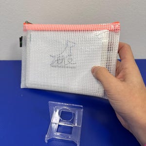 May include: A white mesh zippered pouch with an orange zipper and a small black and white dog logo. The pouch is sitting on a blue surface with a clear plastic stand.