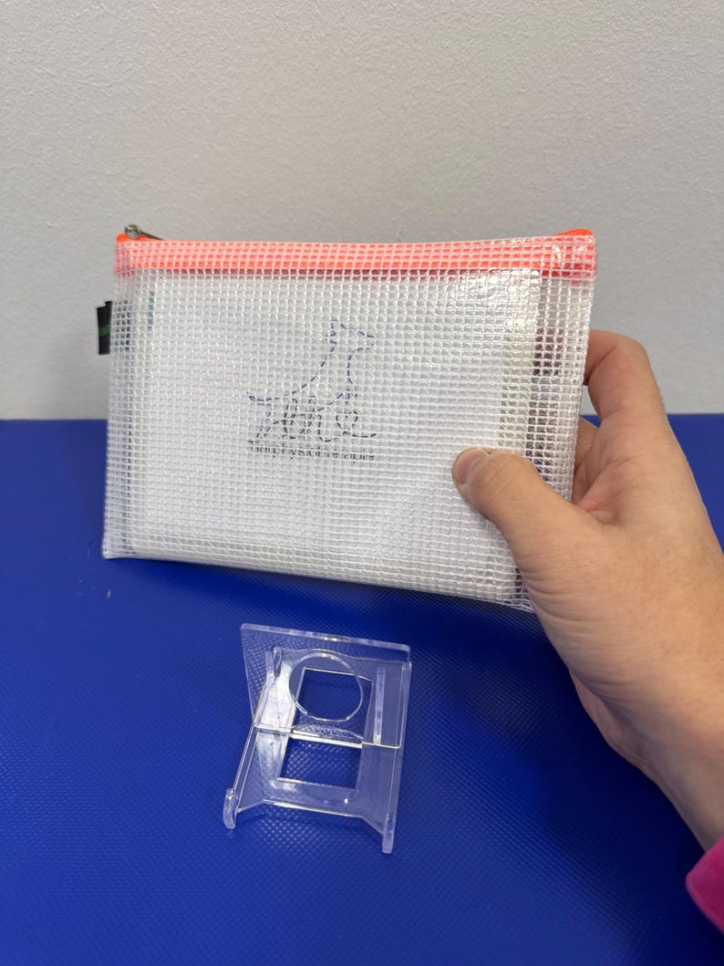 May include: A white mesh zippered pouch with an orange zipper and a small black and white dog logo. The pouch is sitting on a blue surface with a clear plastic stand.