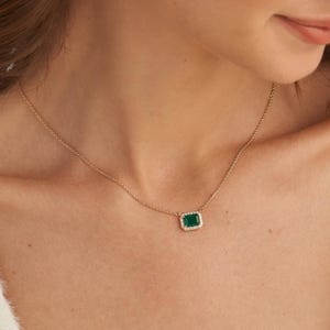 Op de afbeelding: Een gouden ketting met een rechthoekige, smaragdgeslepen groene edelsteen hanger. De centrale groene steen is omringd door een halo van kleine, heldere, fonkelende steentjes, gezet in een gepolijst gouden metalen frame aan een fijne ketting.