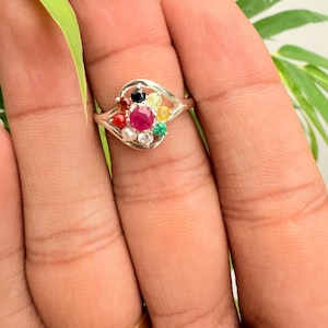 May include: A silver ring featuring a cluster of colorful gemstones, including a central ruby, surrounded by smaller stones in various hues. The ring is held up by a hand, showcasing the intricate design and vibrant colors of the jewelry.