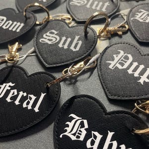 May include: A set of black heart-shaped keychains with different words printed in white gothic font. The words include "Slut", "Dom", "Sub", "Feral", "Baby", "Cunt", "Pup", and "D".