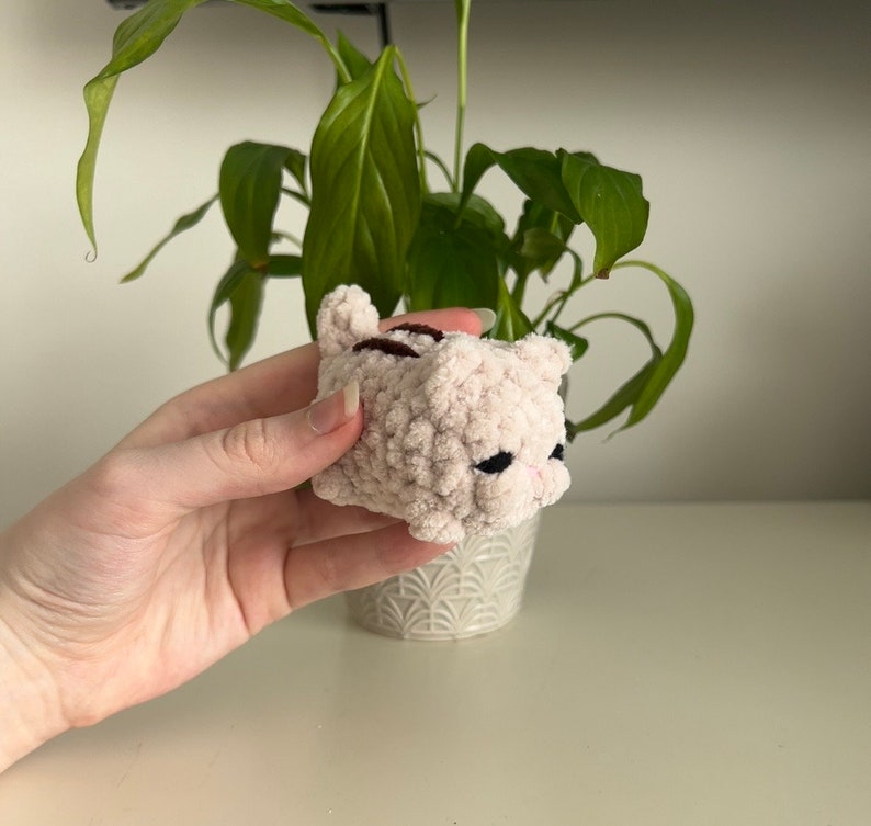May include: A small, pink crocheted cat toy with brown stripes. The cat is lying on its side with its eyes closed.