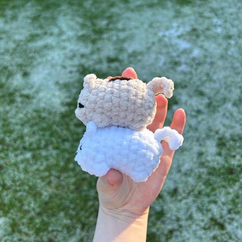 May include: Two crocheted stuffed toys, one white and one beige, are stacked on top of each other. The white toy is on the bottom and the beige toy is on top. The toys are both small and have a rounded shape.