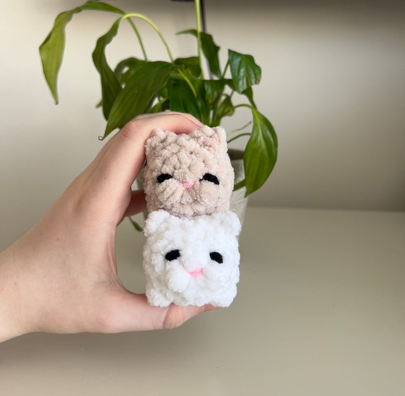 May include: Two crocheted cat toys, one beige and one white, with black eyes and pink noses. The toys are stacked on top of each other and being held by a hand.
