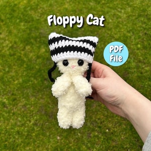 May include: A handmade, fluffy white cat doll with a black and white striped hat and black yarn hair. The doll has large black eyes and a pink nose. The text "Floppy Cat" is above the doll, and a "PDF File" button is visible.