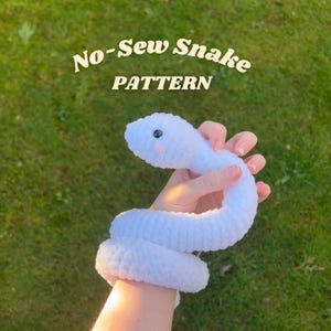 May include: A soft, white crocheted snake toy with a smiling face and black eyes is held against a green grassy background. The text "No-Sew Snake PATTERN" is displayed in a handwritten font above the snake.