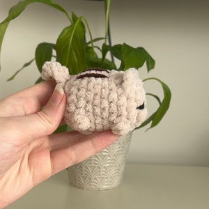 May include: A small, crocheted plush toy shaped like a cat. The cat is light pink with brown stripes and black eyes.