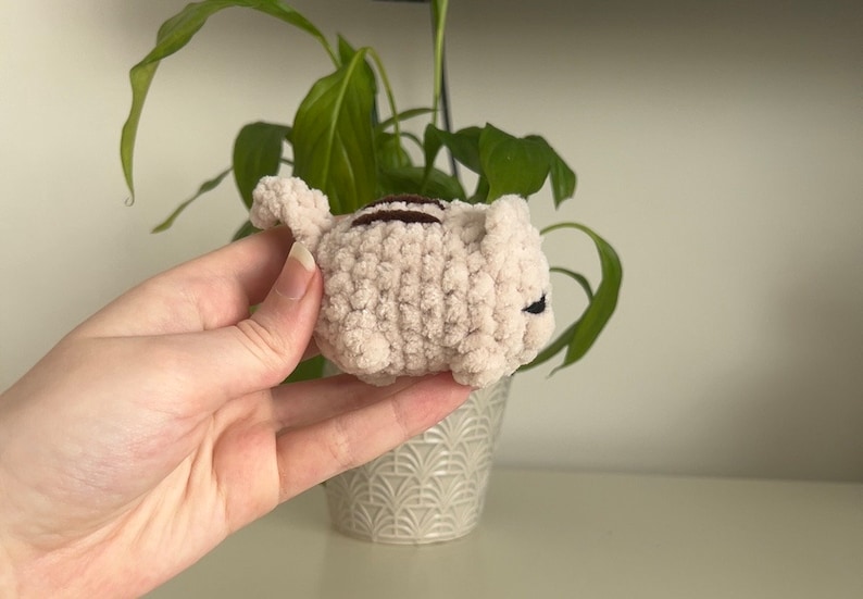 May include: A small, crocheted plush toy shaped like a cat. The cat is light pink with brown stripes and black eyes.