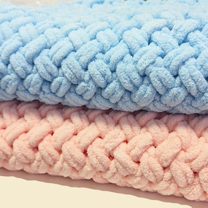 Könnte beinhalten: Zwei gestrickte Decken, eine hellblau und eine rosa, mit einem gewebten Muster. Die Decken sind aus weichem, flauschigem Garn.