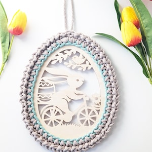 Könnte beinhalten: Osterei-förmige Holzdekoration mit gehäkeltem grauem Rand und hellblauem Akzent. Der Holzausschnitt zeigt einen Hasen auf einem Fahrrad, einen Vogel und ein Osterei. Eine beige Schnur ist zum Aufhängen befestigt.