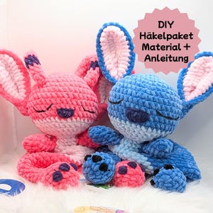 Könnte beinhalten: Zwei gehäkelte Plüschtiere, eines rosa und eines blau, mit geschlossenen Augen und gestickten Details. Die Spielzeuge sitzen auf einer weißen, flauschigen Oberfläche. Der Text "DIY Häkelpaket Material + Anleitung" ist in der oberen rechten Ecke des Bildes sichtbar.