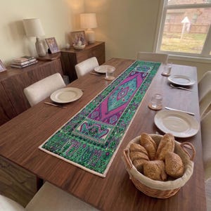 Op de afbeelding: Een rechthoekige tafelloper met een levendig geometrisch patroon in magenta en groenblauw met gedetailleerde randen, plat gelegd op een donkerhouten eettafel met witte borden, glazen en een rieten mand gevuld met broodjes.