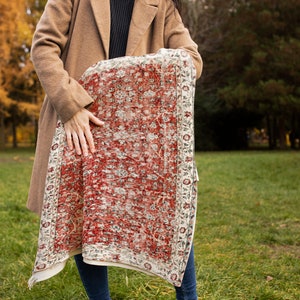 May include: A woman holds a red and white patterned rug in front of her. The rug has a floral design and a white border.
