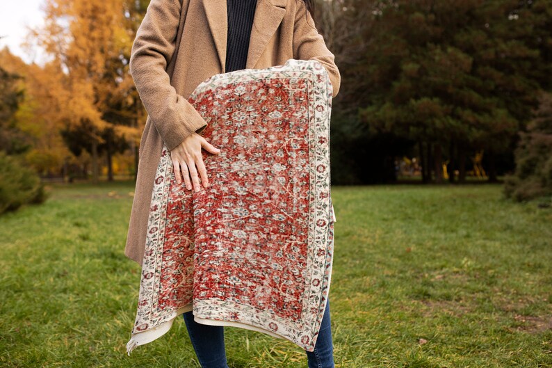 May include: A woman holds a red and white patterned rug in front of her. The rug has a floral design and a white border.