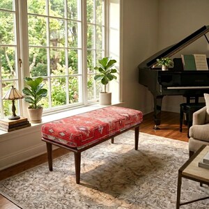 May include: A rectangular bench featuring a red fabric cushion with a geometric pattern and four dark brown tapered wooden legs. The bench is positioned in a room with a large window, a black grand piano, and two potted green plants.