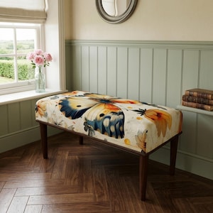 May include: A rectangular upholstered bench featuring a large, detailed butterfly print with orange, blue, and cream tones on the seat. The bench is supported by four dark brown wooden legs and has a cushioned top surface with a floral pattern.