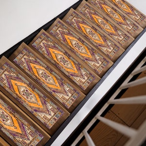 Puede incluir: Un conjunto de peldaños de escalera con patrones en tonos naranja, morado y beige. Los peldaños están hechos de una tela tejida con un diseño geométrico. Los peldaños están instalados en una escalera de madera.