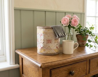 May include: A cylindrical woven storage basket with a cream, yellow, and red geometric pattern, topped with a folded grey and white herringbone fabric. Beside it sits a plain cream ceramic mug, a glass vase with pink roses, and a potted ivy plant.