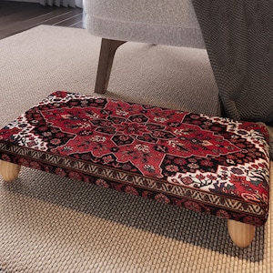 May include: A rectangular footstool with a red, black, and white patterned top, reminiscent of a Persian rug. The footstool has four short, rounded wooden legs. The design features floral and geometric motifs.