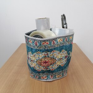 May include: A teal and white patterned fabric storage basket with a roll of clear packing tape, a roll of brown packing tape, and a silver tool inside.