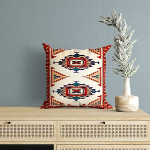 May include: A decorative throw pillow with a geometric pattern in shades of red, blue, and white. The pillow has a cream-colored background and a border of red and white.