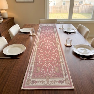 May include: A rectangular table runner featuring a dusty pink center panel with a symmetrical white scrollwork pattern, bordered by a cream-colored frame decorated with intricate floral motifs and leaf designs along the edges.