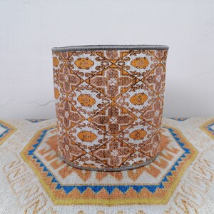 May include: A cylindrical fabric storage bin with a geometric pattern in shades of brown, white, and yellow. The bin has a gray trim around the top.