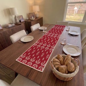May include: A rectangular red table runner featuring a repeating white snowflake pattern, centered on a dark wood dining table. The table is set with white plates, glasses, and a woven basket filled with various crusty bread rolls.