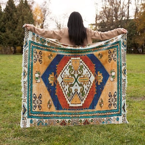 May include: A woman holds a colorful patterned throw blanket with a geometric design in shades of blue, orange, yellow, and green. The blanket has a fringed edge and is held out in front of her.