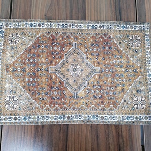 Könnte beinhalten: Ein rechteckiger brauner, blauer und weißer Teppich mit geometrischem Muster. Der Teppich hat einen Rand mit einem sich wiederholenden Muster aus kleinen Quadraten.