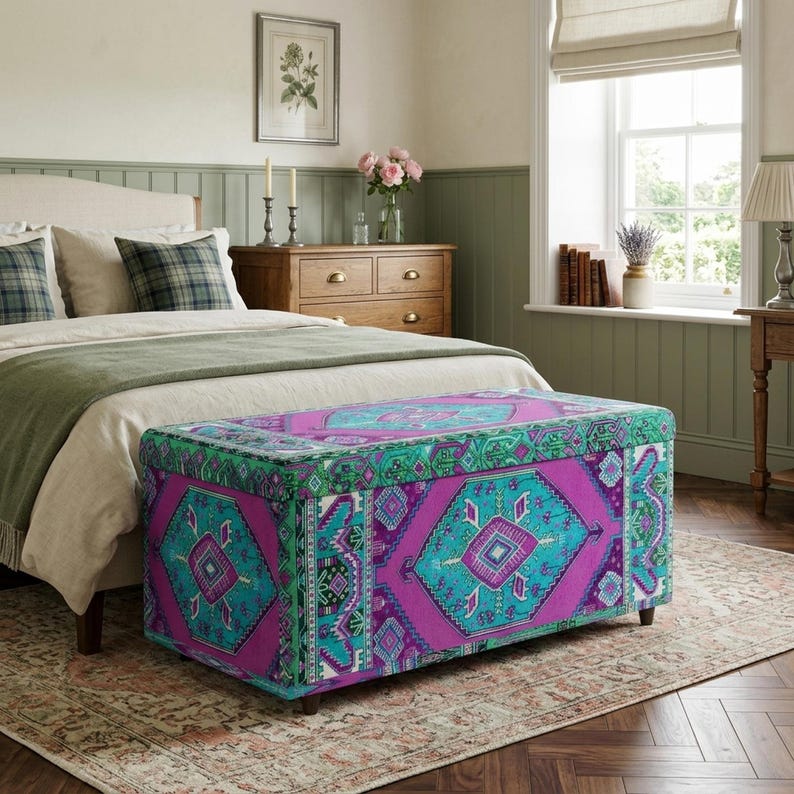 a cozy bedroom with a large bed, a wooden dresser, and a colorful patterned ottoman. The room has a warm and inviting atmosphere, with a window providing natural light and a vase of flowers on the dresser.