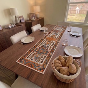 Op de afbeelding: Een rechthoekige tafelloper met een gedetailleerd geometrisch patroon in oranje, grijs, wit en zwart, geplaatst op een donkerhouten eettafel. De loper is omringd door witte borden, glazen en een rieten mand gevuld met verschillende soorten brood.