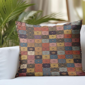 Puede incluir: Cojín cuadrado con un patrón geométrico colorido. El diseño presenta una cuadrícula de cuadrados, cada uno con un motivo floral en tonos naranja, azul, amarillo y rosa. El cojín está colocado sobre un sofá blanco.