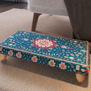 May include: A rectangular footstool with a teal floral pattern and wooden legs. The pattern features red and white floral designs on a teal background. The footstool is placed on a neutral-coloured rug.