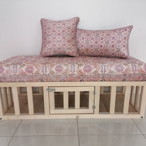 Puede incluir: Un banco de madera con un cojín estampado en rosa y blanco y dos almohadas a juego. El banco tiene un compartimento de almacenamiento integrado con una puerta.