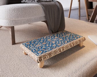 May include: A rectangular footstool with a blue and gold floral pattern. The footstool has a beige border and is supported by four short, light-colored wooden legs. The footstool is placed on a beige rug.