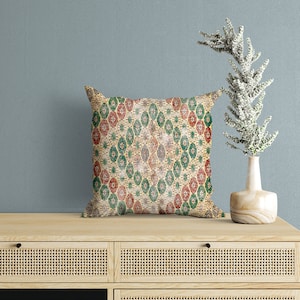May include: A decorative throw pillow with a geometric pattern in shades of green, red, and beige. The pillow is sitting on a wooden shelf with a vase of white flowers.