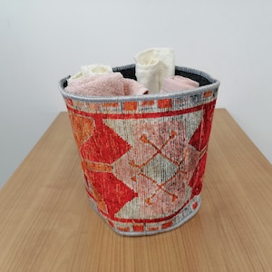 May include: A red, orange, and white patterned fabric storage basket with a gray trim. The basket is filled with white and pink towels.