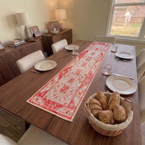 Op de afbeelding: Een lange rechthoekige tafelloper met een levendig rood en crèmekleurig geometrisch patroon op een donkerhouten eettafel. De tafel is gedekt met witte borden, heldere glazen en een gevlochten mand gevuld met knapperige broodjes.