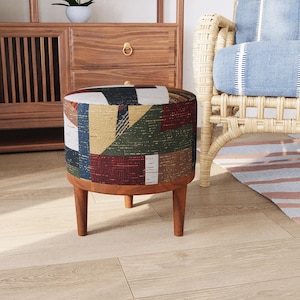 May include: A round ottoman featuring a multi-colored geometric fabric upholstery with shades of red, green, blue, and white. The piece is supported by three tapered wooden legs and includes a matching wooden base frame beneath the seat cushion.