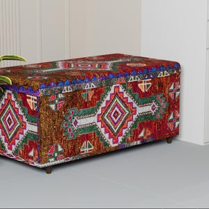 May include: A rectangular storage ottoman with a colorful, patterned fabric. The design features geometric shapes in red, green, white, and blue on a brown background. The ottoman has short, dark wooden legs.